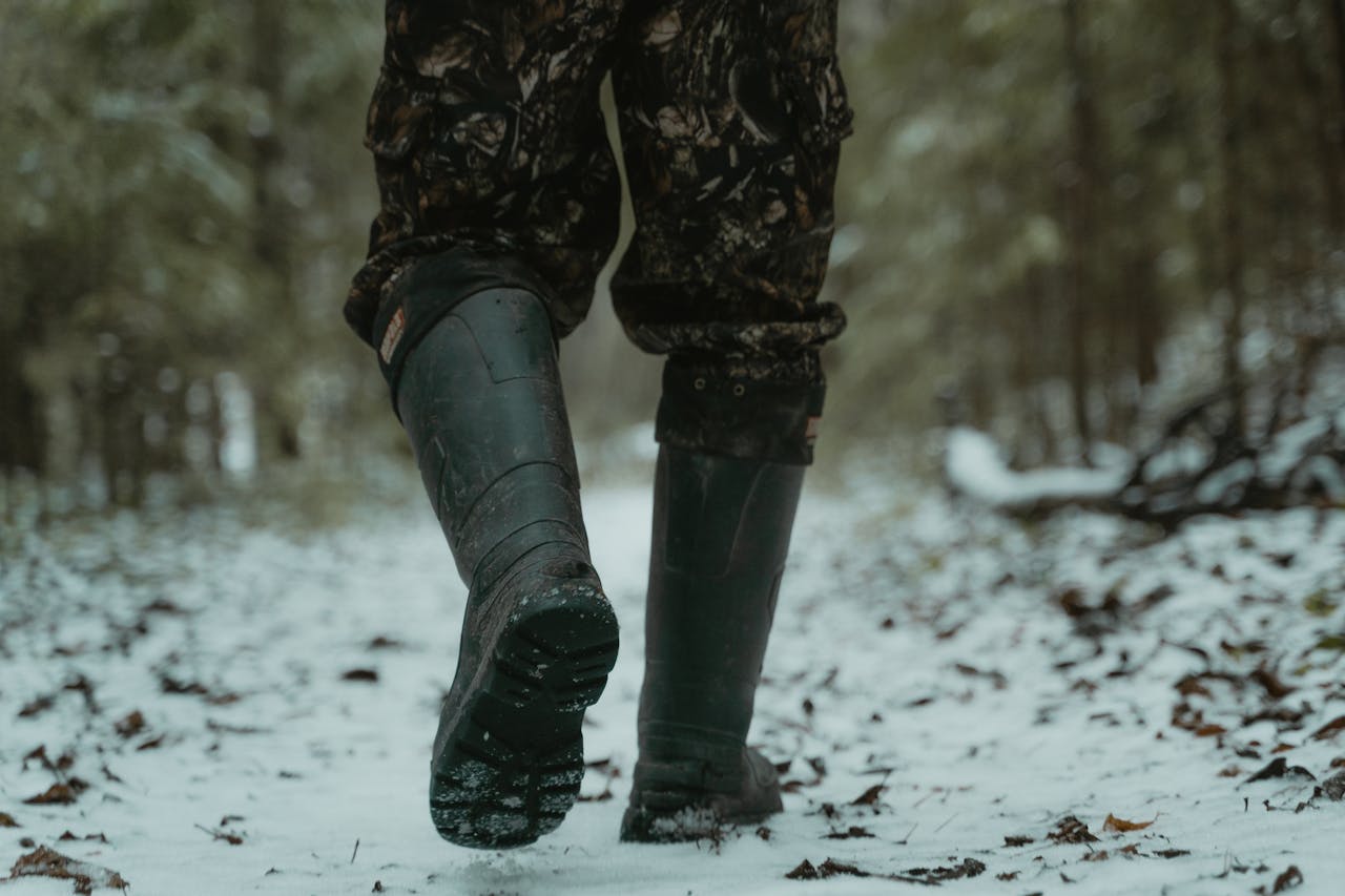 A person in camo pants walking through a snow-covered forest trail, wearing sturdy boots.